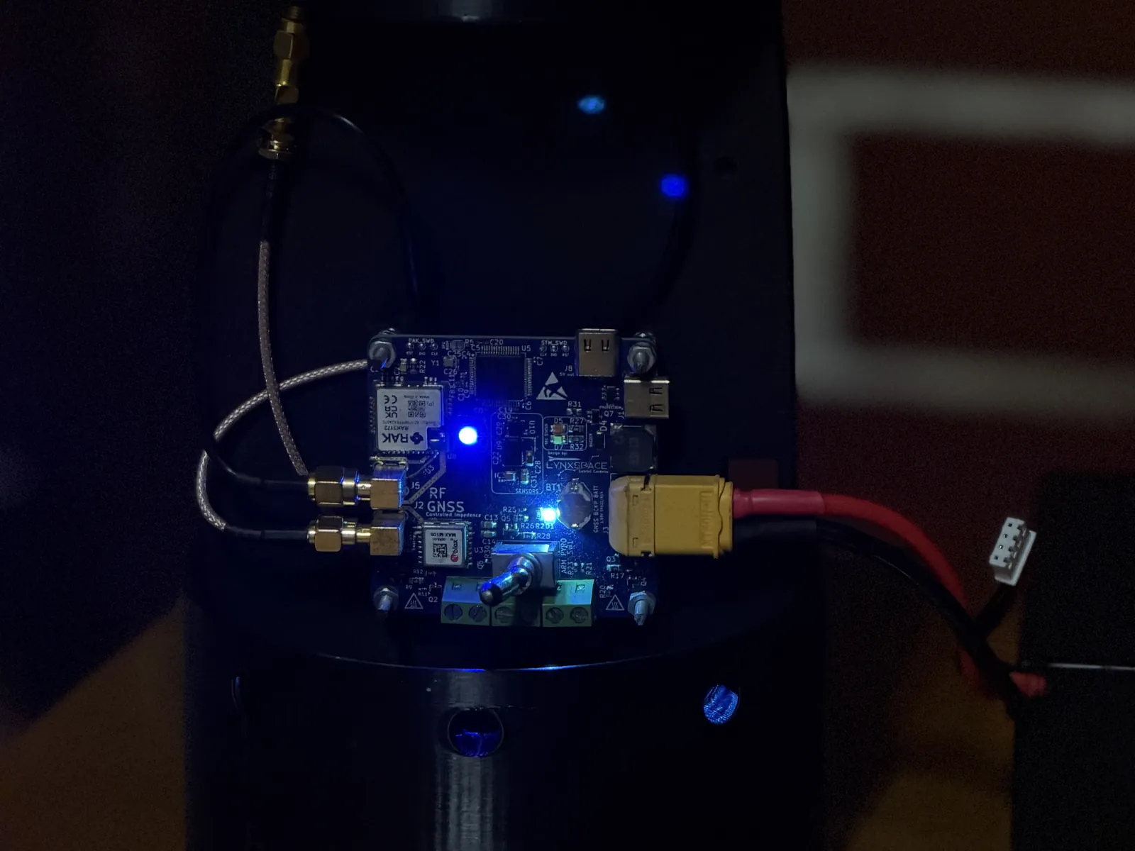 LynxYNav V6.1 powered up on its avionics sled inside the airframe coupler: blue status LEDs lit, XT60 power lead connected, two SMA antenna leads and the LoRa antenna above